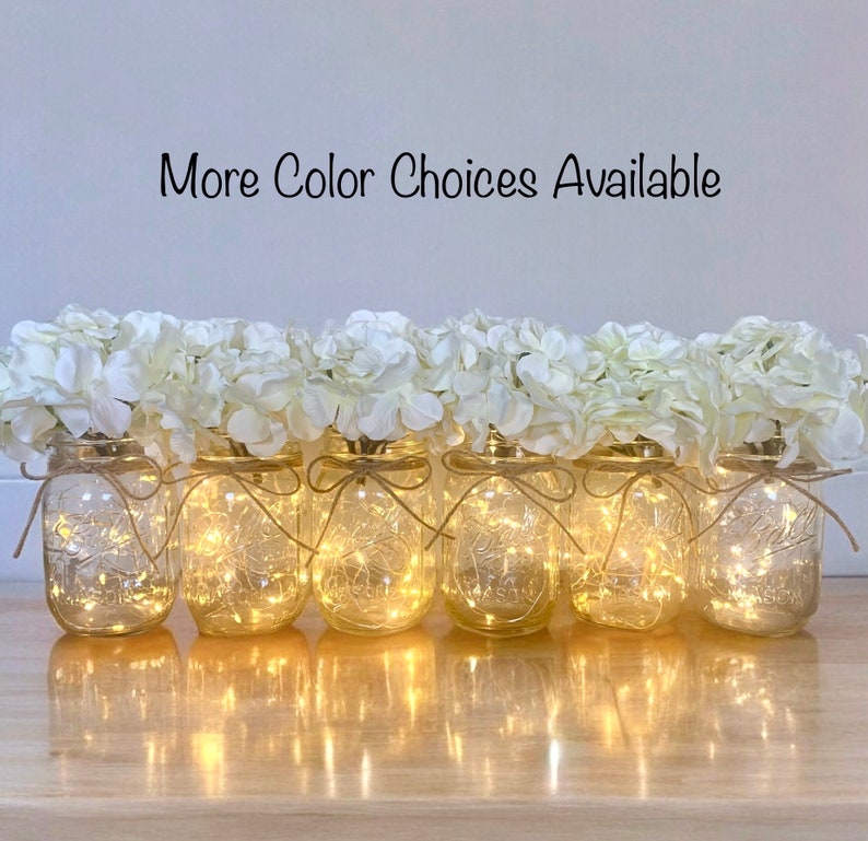 May include: Six clear glass jars with white hydrangea flowers and twinkling fairy lights. The jars are tied with twine and the text "More Color Choices Available" is at the top of the image.