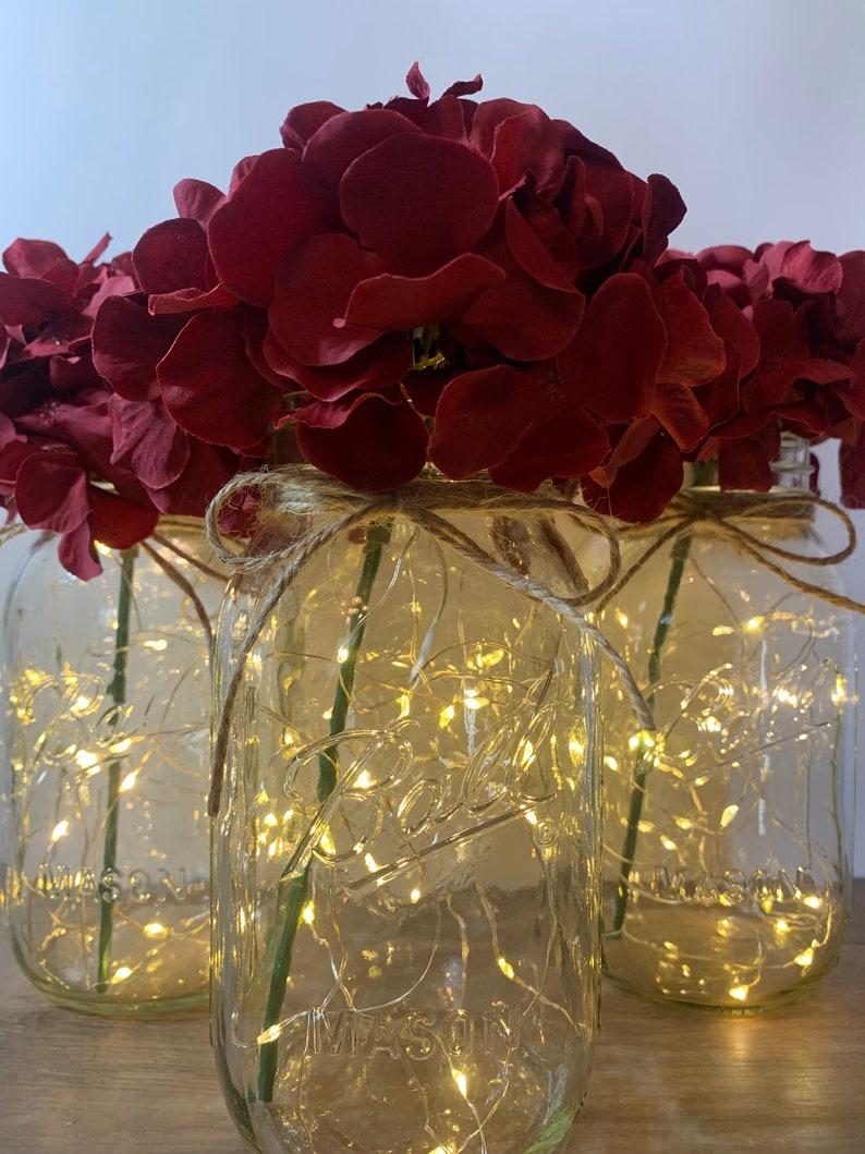 May include: Three clear glass mason jars with burgundy flowers and white fairy lights inside. The jars are tied with twine around the top.