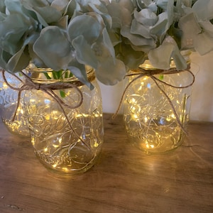 May include: Three clear glass mason jars with a rustic twine bow around the top. Each jar contains a string of fairy lights and is filled with light blue artificial hydrangeas.