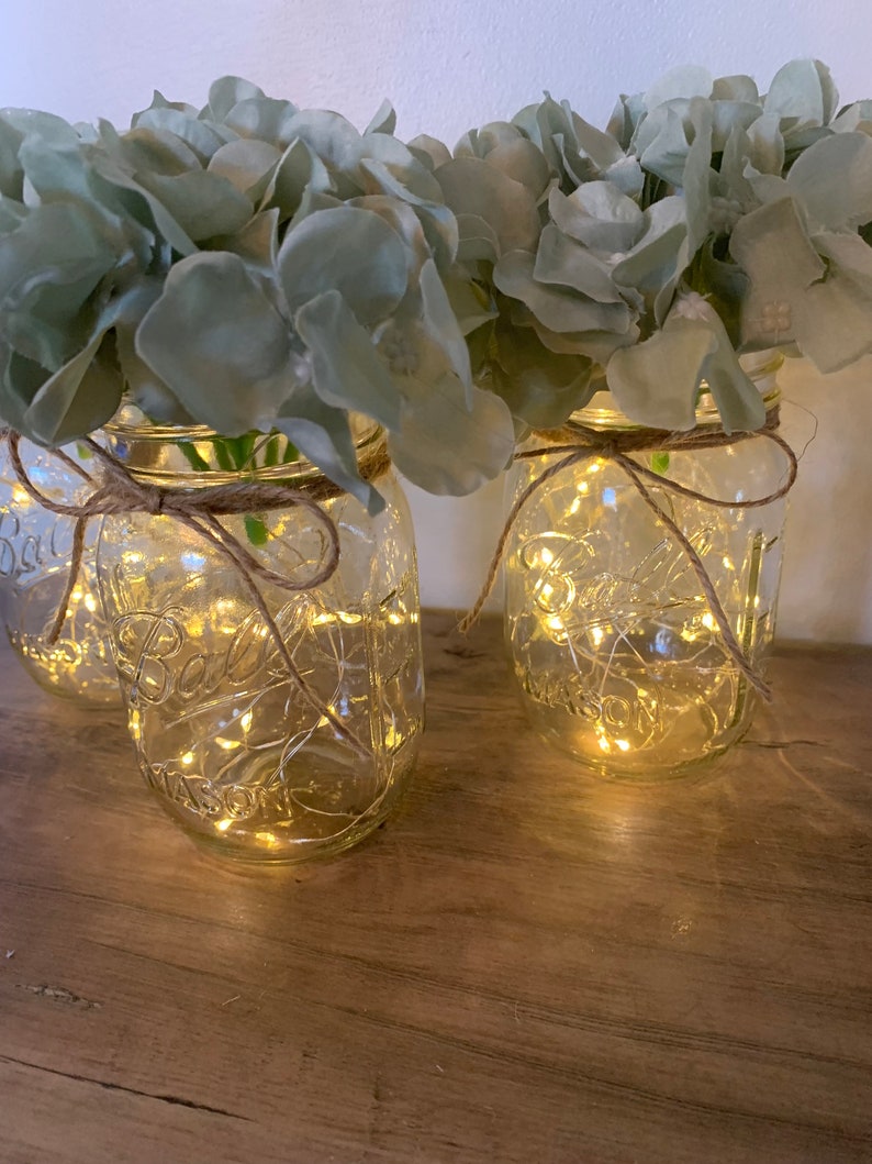 May include: Three clear glass mason jars with a rustic twine bow around the top. Each jar contains a string of fairy lights and is filled with light blue artificial hydrangeas.