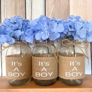 K&ouml;nnte beinhalten: Drei Glasvasen mit hellblauen Hortensienbl&uuml;ten, jede mit einem Juteband und dem Text "It's a Boy" auf der Vorderseite.