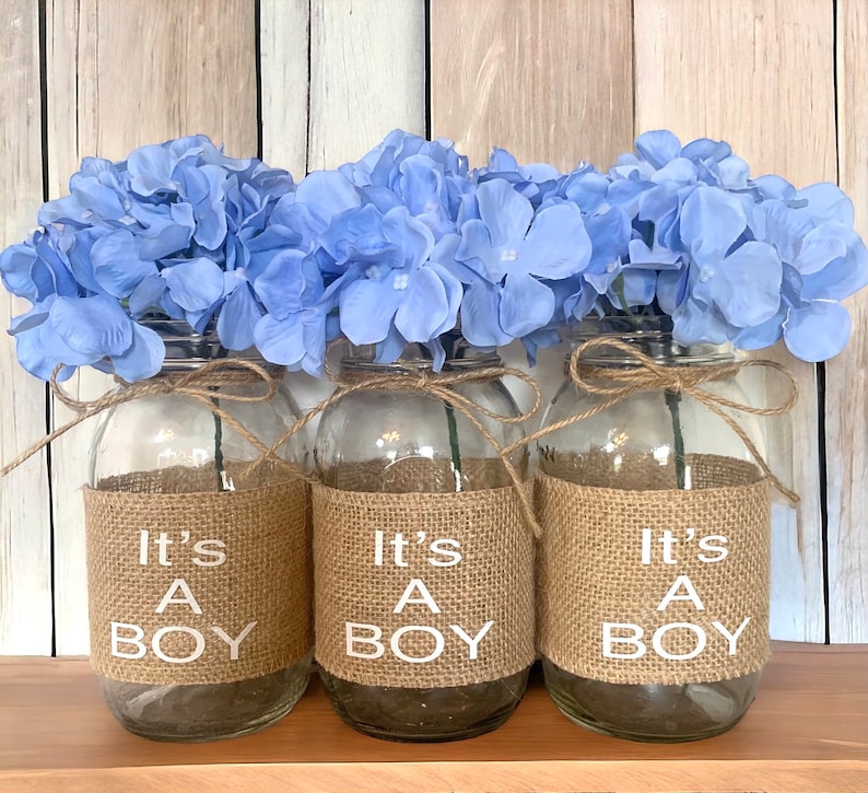 K&ouml;nnte beinhalten: Drei Glasvasen mit hellblauen Hortensienbl&uuml;ten, jede mit einem Juteband und dem Text "It's a Boy" auf der Vorderseite.