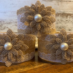 May include: Three burlap napkin rings with white lace trim and a large brown burlap flower with a pearl center.