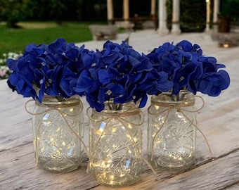 May include: Three clear glass mason jars with blue hydrangea flowers and string lights inside. The jars are tied with twine and are sitting on a wooden table.