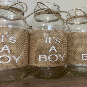 K&ouml;nnte beinhalten: Drei durchsichtige Glasbeh&auml;lter mit Jutes&auml;cken, auf denen in wei&szlig;en Buchstaben "It's A Boy" steht. Die Beh&auml;lter sind mit Schnur gebunden.