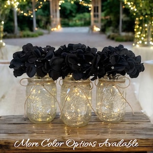 May include: Three clear glass mason jars with black hydrangea flowers and string lights inside. The jars are sitting on a wooden surface.