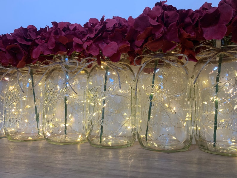 May include: A row of clear glass mason jars with burgundy flowers and white fairy lights inside. The jars are tied with twine and are sitting on a wooden surface.