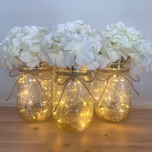 May include: Three clear glass mason jars with white hydrangea flowers and string lights inside. The jars are tied with twine and bows.