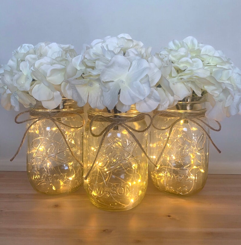 May include: Three clear glass mason jars with white hydrangea flowers and string lights inside. The jars are tied with twine and bows.