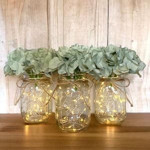 May include: Three clear glass mason jars with a rustic twine bow around the top. Each jar is filled with faux greenery and a string of fairy lights.