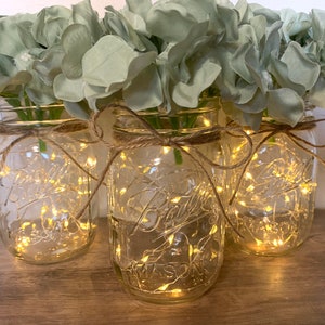 May include: Three clear glass mason jars with a string of fairy lights inside, decorated with burlap ribbon and filled with light green hydrangea flowers.