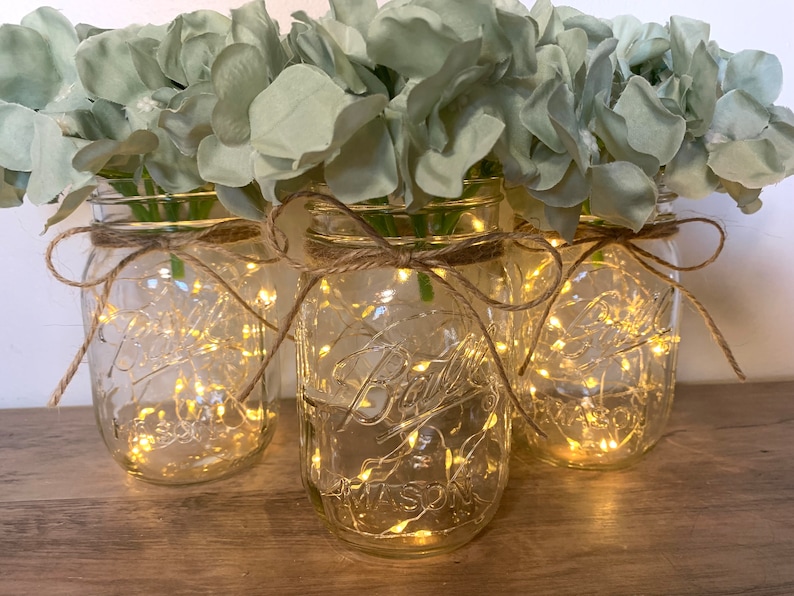May include: Three clear glass mason jars with a string of fairy lights inside, decorated with burlap ribbon and filled with light green hydrangea flowers.