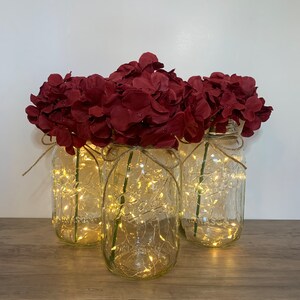 May include: Three clear glass jars with a string of fairy lights inside, each filled with a bouquet of burgundy artificial hydrangeas.
