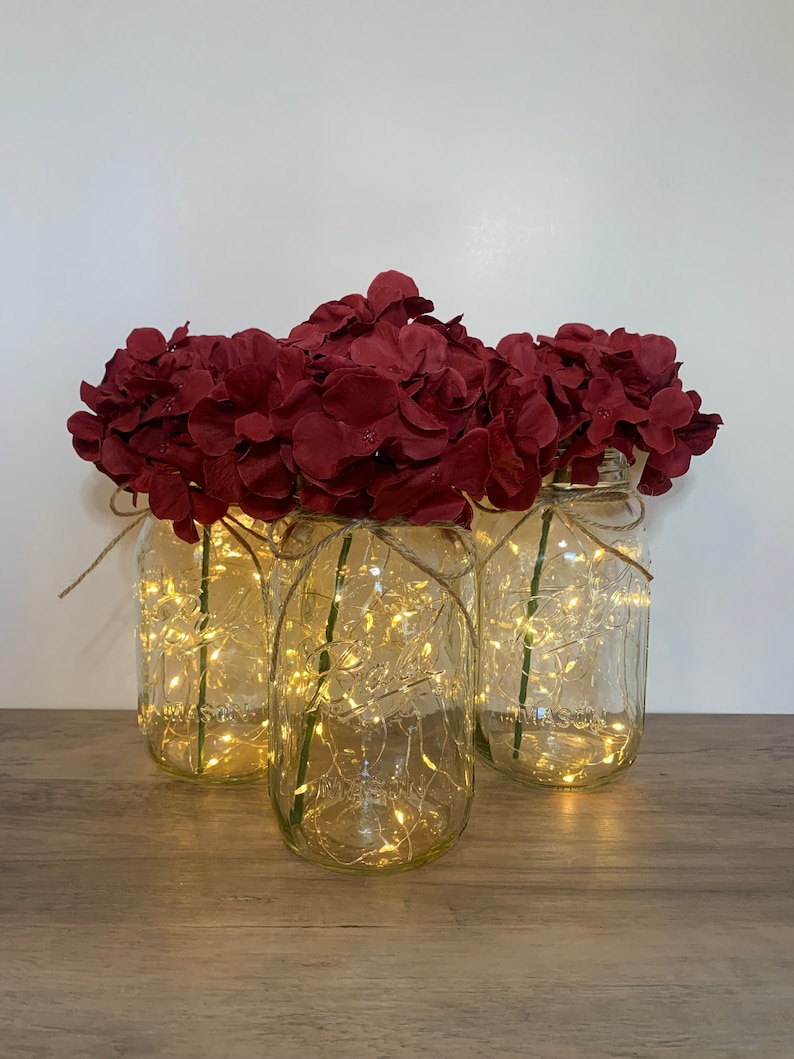 May include: Three clear glass jars with a string of fairy lights inside, each filled with a bouquet of burgundy artificial hydrangeas.