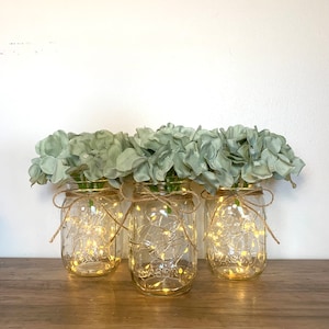May include: Three clear glass mason jars with a string of fairy lights inside each jar. The jars are tied with twine and decorated with light green artificial hydrangea flowers.
