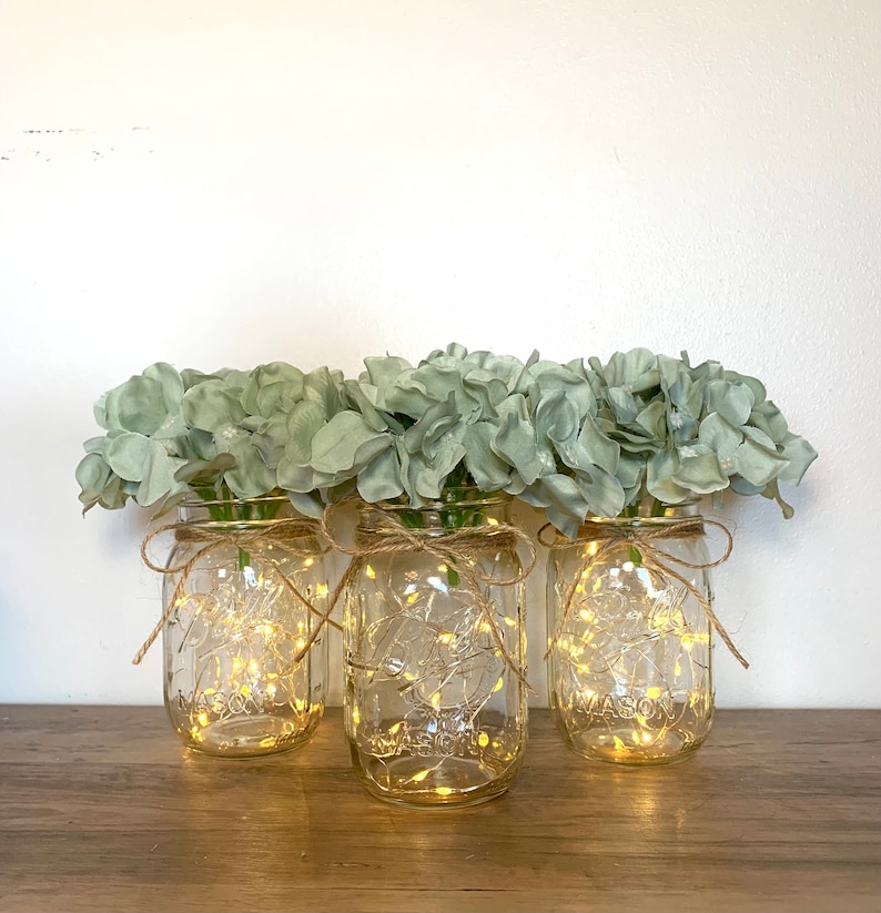 May include: Three clear glass mason jars with a string of fairy lights inside each jar. The jars are tied with twine and decorated with light green artificial hydrangea flowers.