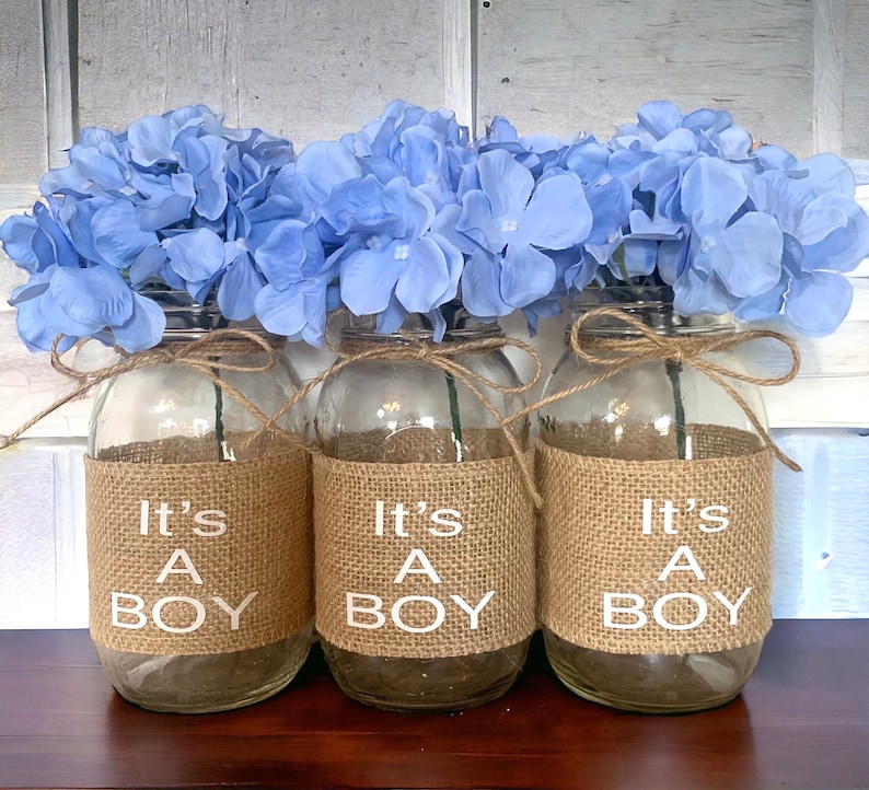K&ouml;nnte beinhalten: Drei Glasgl&auml;ser mit Juteband und einem Juteband um die Mitte. Jedes Glas hat ein wei&szlig;es Etikett mit der Aufschrift "It's A Boy". Die Gl&auml;ser sind mit hellblauen Hortensien gef&uuml;llt.