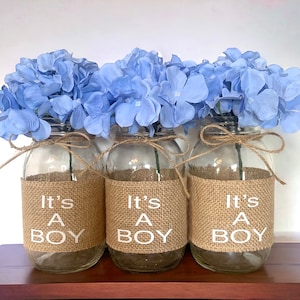K&ouml;nnte beinhalten: Drei Glasvasen mit einem Jutestoffband und dem Text "It's A Boy" in wei&szlig;en Buchstaben. Jede Vase ist mit hellblauen Hortensien gef&uuml;llt.