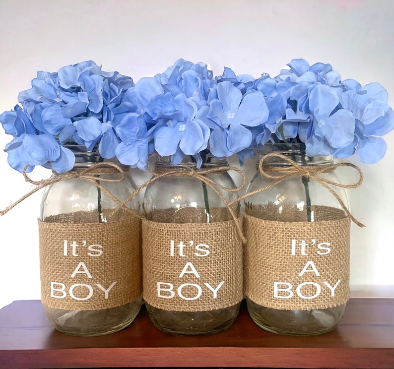 K&ouml;nnte beinhalten: Drei Glasvasen mit einem Jutestoffband und dem Text "It's A Boy" in wei&szlig;en Buchstaben. Jede Vase ist mit hellblauen Hortensien gef&uuml;llt.