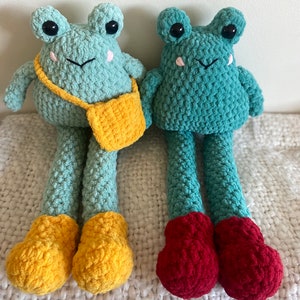 May include: Two crocheted frog plushies with long legs. The frog on the left is wearing yellow shoes and a yellow bag. The frog on the right is wearing red shoes.