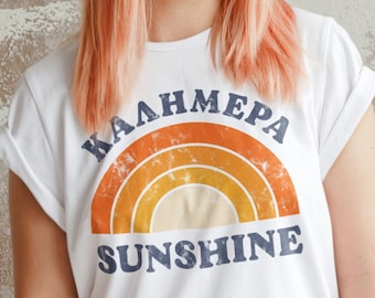 Peut inclure: Un t-shirt blanc avec un graphique arc-en-ciel rétro et le texte "Kalimera Sunshine".