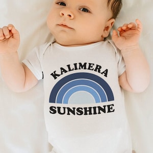 May include: A white baby onesie with the text "Kalimera Sunshine" in a blue rainbow design. The baby is lying on a white sheet.