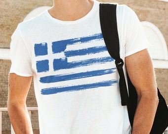 May include: White t-shirt featuring a blue and white Greek flag design. The flag is painted with brushstrokes. The shirt is worn with blue jeans and sunglasses. A black backpack is worn over the shoulder.