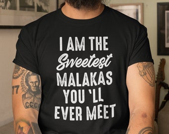 May include: A man wearing a black t-shirt with the text "I am the Sweetest Malakas You'll Ever Meet" in white. He is wearing a black beanie and has a beard. He is sitting on a white couch in a room with framed pictures on the walls.