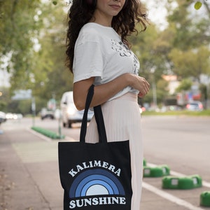 Peut inclure: Une femme portant un t-shirt blanc et une jupe rose porte un sac fourre-tout noir avec un arc-en-ciel bleu et le texte "Kalimera Sunshine".
