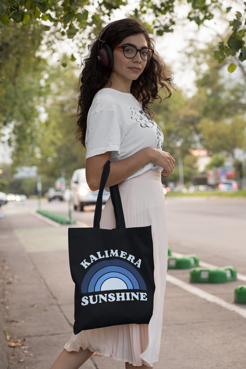 Peut inclure: Une femme portant un t-shirt blanc et une jupe rose porte un sac fourre-tout noir avec un arc-en-ciel bleu et le texte "Kalimera Sunshine".