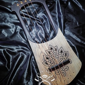 May include: A wooden lyre with a dark frame and a light wood soundboard. The soundboard features a black Celtic knot design. The instrument has six strings and gold tuning pegs. The lyre rests on black satin fabric. The instrument is approximately 60 cm in length.