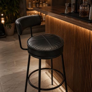 May include: A black bar stool featuring a circular, padded seat with cross-stitched detailing and a curved backrest. The stool is supported by a black metal frame with four straight legs and a circular footrest ring connecting the legs for stability.