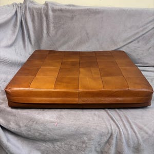 May include: A brown leather square ottoman with a quilted design. The ottoman is sitting on a gray blanket.