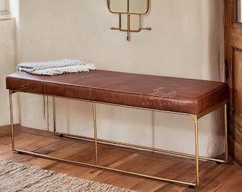 May include: A brown leather bench with a rectangular seat and gold-colored metal legs. A folded blanket rests on the bench. The bench is in a room with a wooden floor and a mirror on the wall. The words "Epic Leather" are printed on the bench.