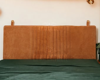 Könnte beinhalten: Ein braunes Lederkopfteil mit vertikalen Nähten und goldenen Metallhaken. Das Kopfteil ist an einem Bett mit einem dunkelgrünen Bettbezug befestigt.