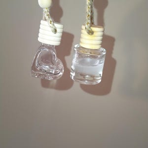 Op de afbeelding: Twee glazen autoluchtverfrissers die aan een touwtje hangen. Eén is een fles in de vorm van een hart en de andere is een ronde fles. Beide flessen zijn helder en gevuld met een vloeistof.