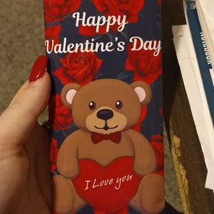 Puede incluir: Una tarjeta de San Valentín Swisher Sweets roja, blanca y azul con un osito de peluche que sostiene un corazón rojo que dice "Te amo". La tarjeta tiene un fondo de rosas rojas sobre un fondo azul oscuro.