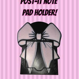 Op de afbeelding: Een zwarte Post-it notitieblokhouder met een groot strikdesign. De strik is wit met roze schaduwen en zwarte contouren. De tekst "POST-IT NOTE PAD HOLDER!" staat boven de houder op een roze gestreepte achtergrond.