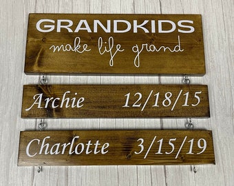 May include: A wooden sign with the text "GRANDKIDS make life grand" on the top, followed by two lines with names and dates. The top line reads "Archie 12/18/15" and the bottom line reads "Charlotte 3/15/19".