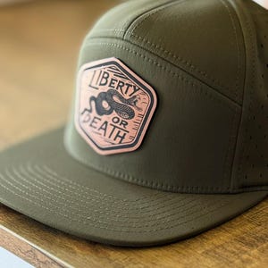 May include: Olive green baseball cap with a flat brim. The cap features a leather patch with the words "LIBERTY OR DEATH" surrounding a coiled snake illustration. The cap has a perforated design for breathability.