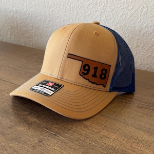 May include: A tan and blue trucker hat with a brown leather patch featuring the outline of the state of Oklahoma and the number 918.