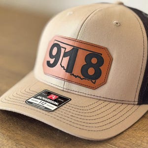 May include: A tan and black trucker hat with a brown leather patch that says "918" on it. The patch is shaped like the state of Oklahoma. The hat is made by Richardson 112.
