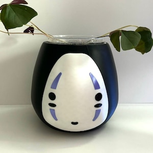 Puede incluir: Taza de cerámica negra con un personaje blanco de No Face de la película El viaje de Chihiro. La taza tiene un contorno morado alrededor de los ojos y la boca. Una planta verde está en el fondo.