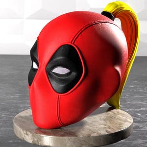 Könnte beinhalten: Eine rote und schwarze Deadpool-Maske mit einem gelben Pferdeschwanz. Die Maske steht auf einem grauen Marmorsockel.