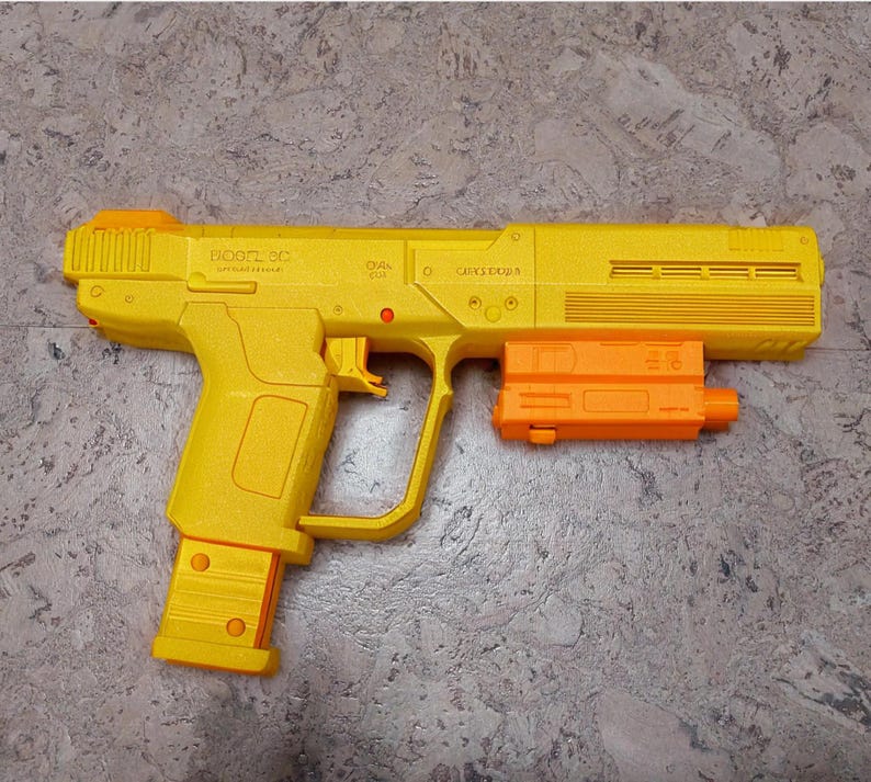 Pu&ograve; includere: Una pistola giocattolo di plastica gialla con un caricatore nero e una canna arancione. La pistola ha il testo "DOOFTL.CC" e "CAPACIDAD 6"