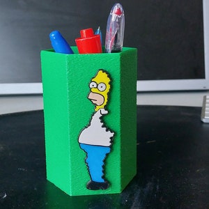 May include: Green hexagonal pencil holder with a cartoon image of Homer Simpson on the front. The holder has three pens inside.