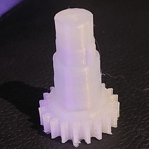 Puede incluir: Un engranaje de pl&aacute;stico blanco con una parte superior cil&iacute;ndrica lisa. El engranaje tiene una serie de dientes alrededor del borde inferior.