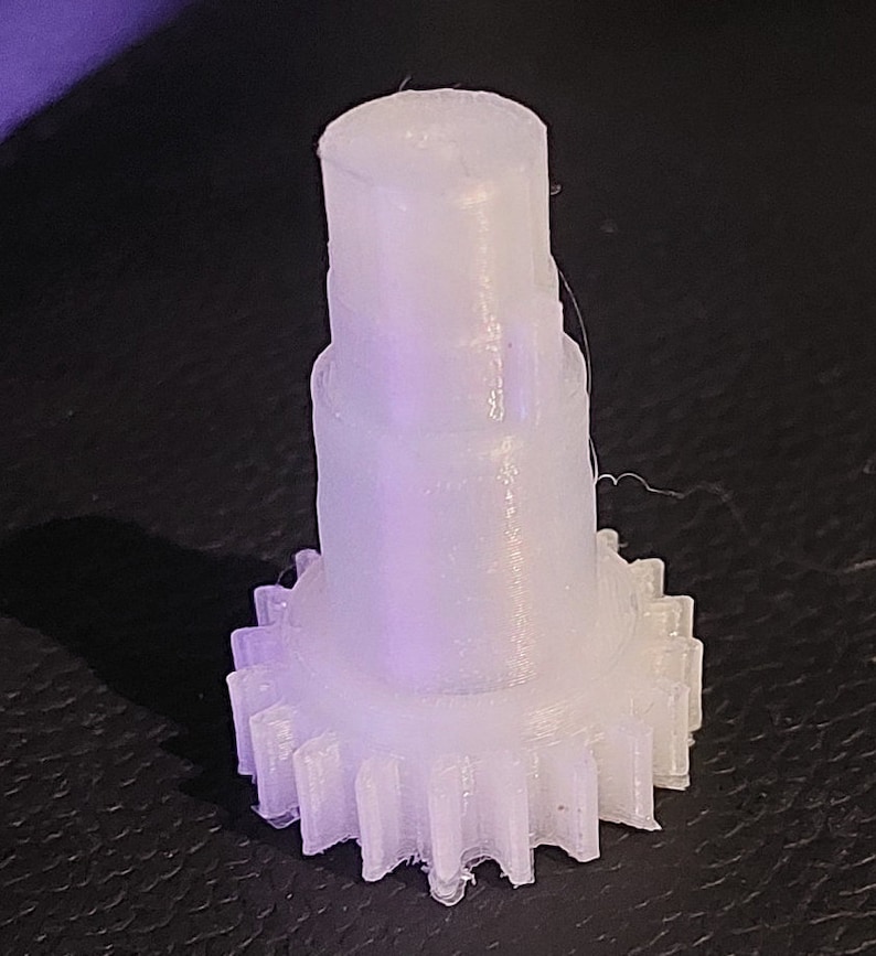 Puede incluir: Un engranaje de pl&aacute;stico blanco con una parte superior cil&iacute;ndrica lisa. El engranaje tiene una serie de dientes alrededor del borde inferior.