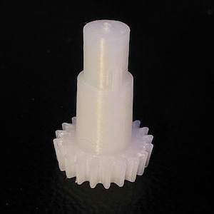 May include: A white plastic gear with a central shaft. The gear has a smooth, rounded top and a toothed bottom.