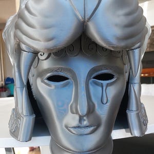 Op de afbeelding: Een zilveren 3D-geprinte helm met een leeuwenkopontwerp. De helm heeft een menselijk gezicht met een traan die langs de wang loopt.
